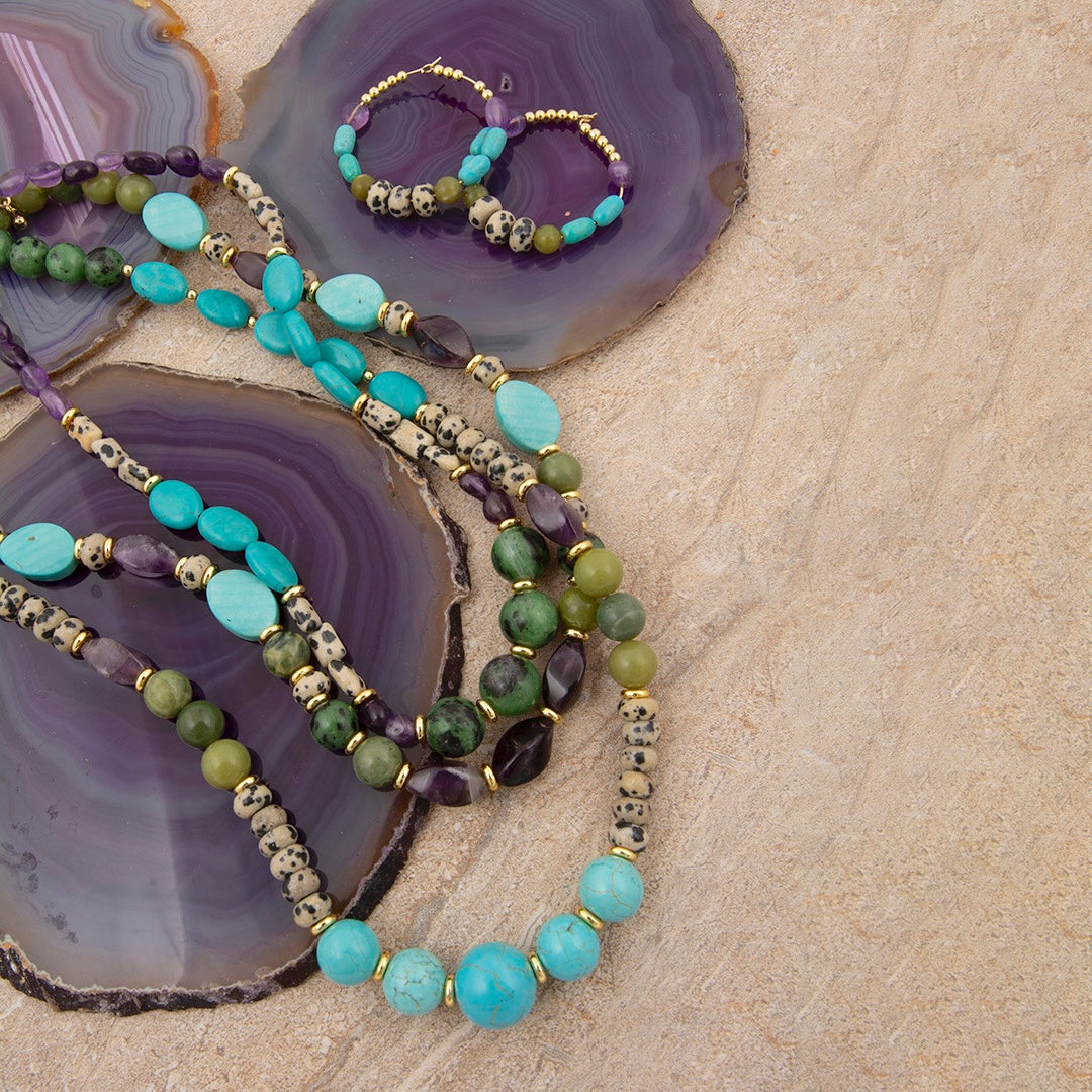 A beaded strand necklace and matching hoop earrings displayed on polished purple agate slices over a beige stone surface. The jewelry features turquoise-blue beads, olive green beads, purple amethyst beads, and gold-toned spacer beads.