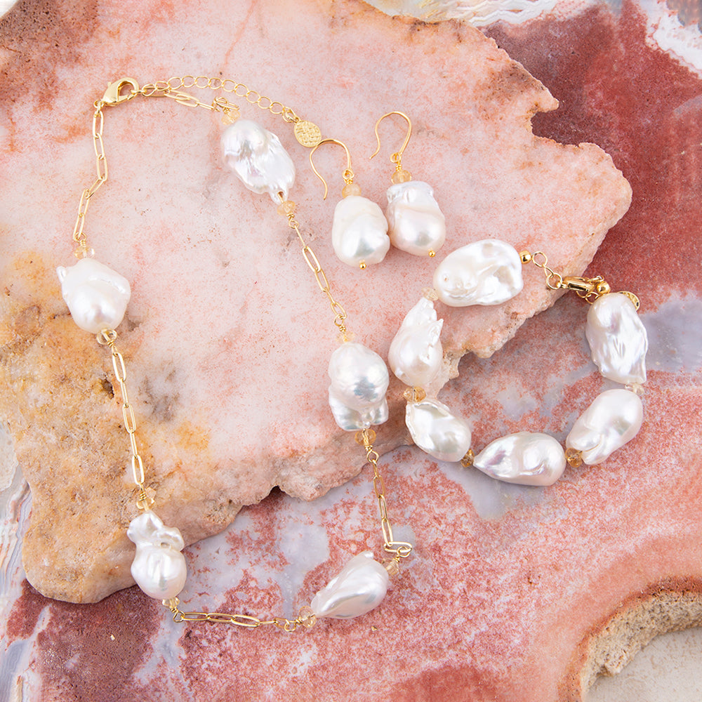 Gold-tone jewelry set featuring a necklace, bracelet, and drop earrings with irregular white baroque pearls, displayed on a pinkish stone surface.
