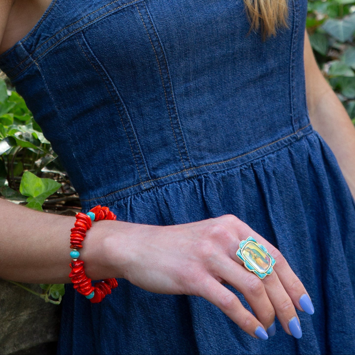 Person wearing a blue dress with a red bracelet and colorful ring, standing against a natural background.