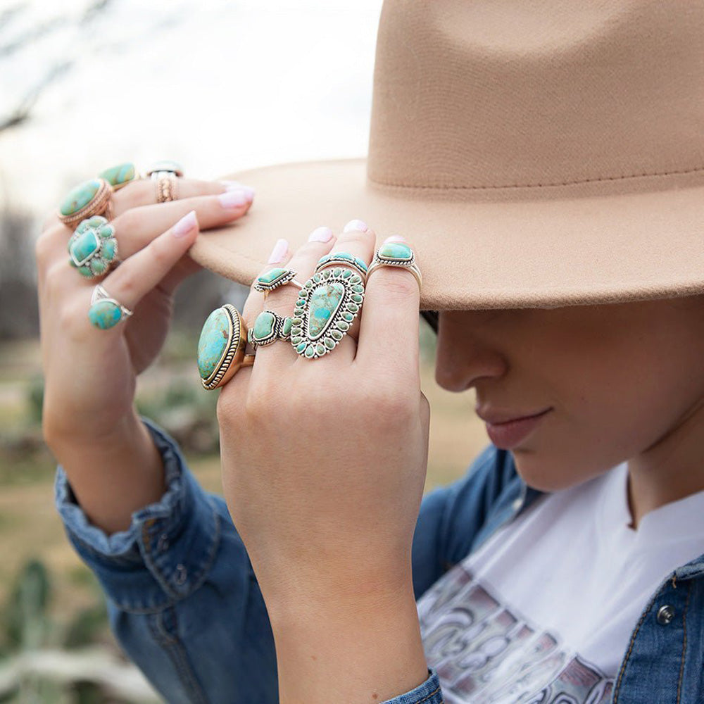 Person wearing multiple turquoise rings with a beige hat and denim jacket.