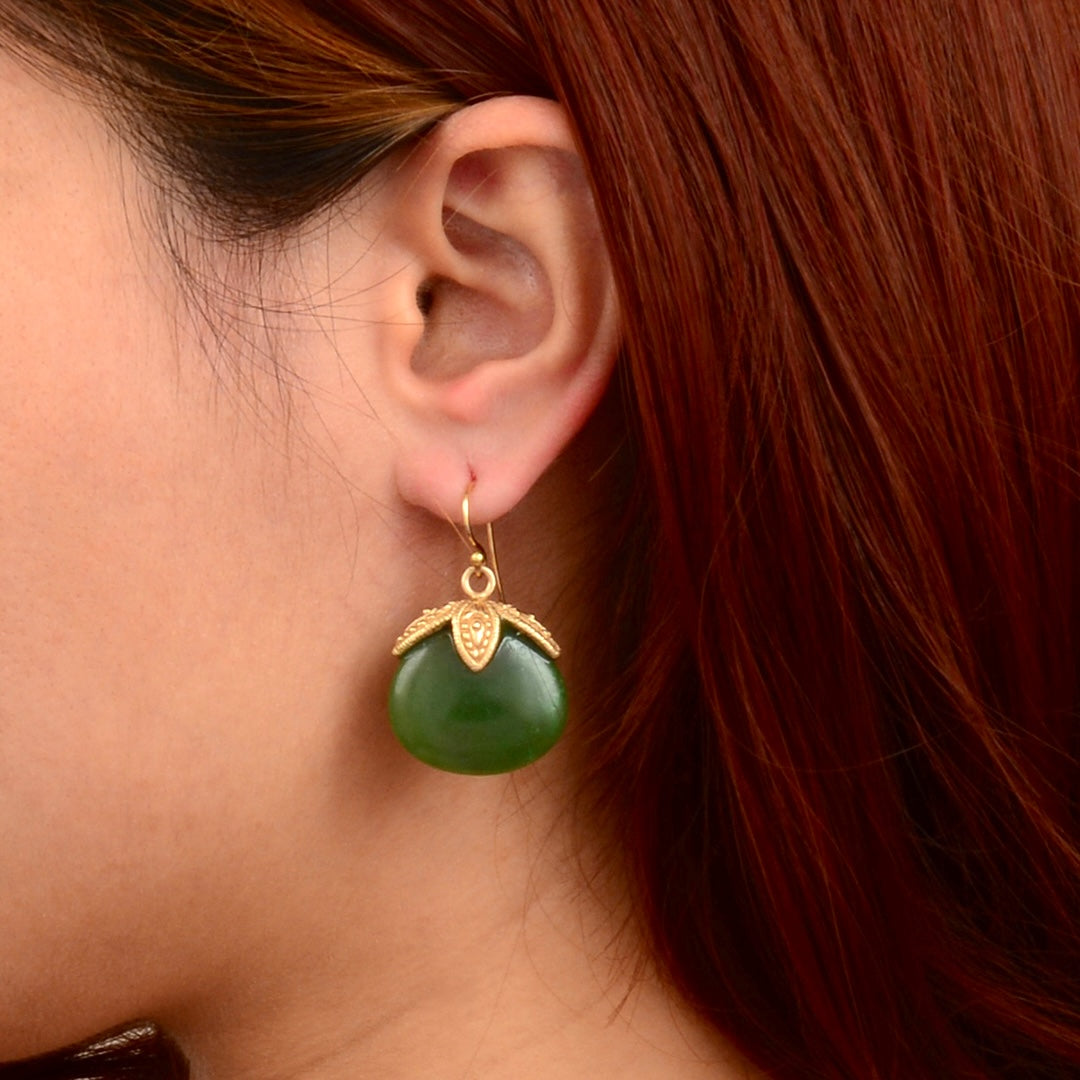 A close-up of a woman's ear wearing a single dangling earring. The earring has a polished green oval stone pendant with a gold-toned decorative top and hook.