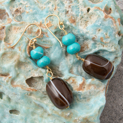 A pair of handmade dangle earrings with gold fishhook wires, three round turquoise beads, and a large polished dark brown agate drop, displayed on a textured turquoise rock.