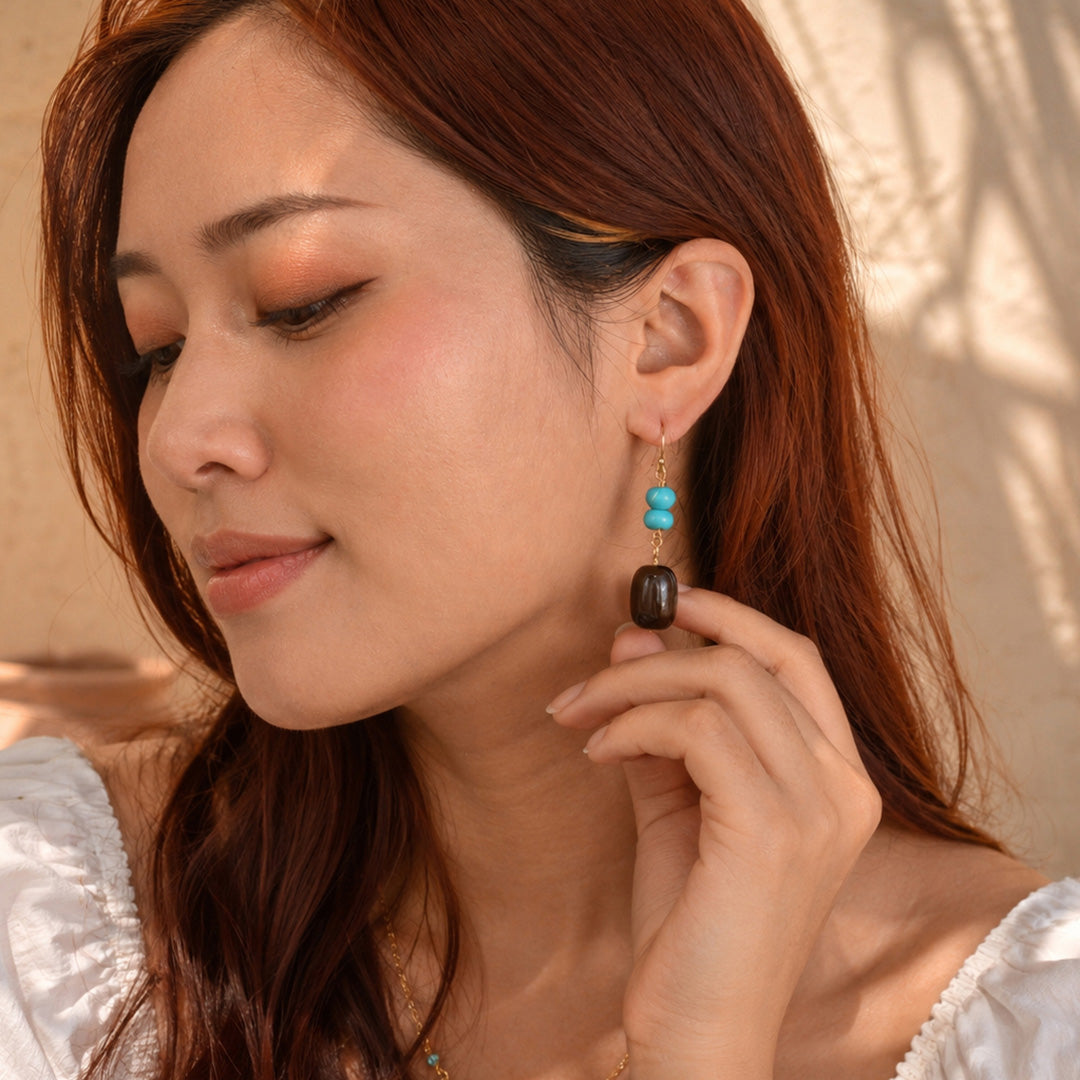 A woman with long reddish-brown hair wearing a dangling earring with three stacked turquoise-blue beads and a dark brown oval charm, posed against a warm beige wall with soft shadows.