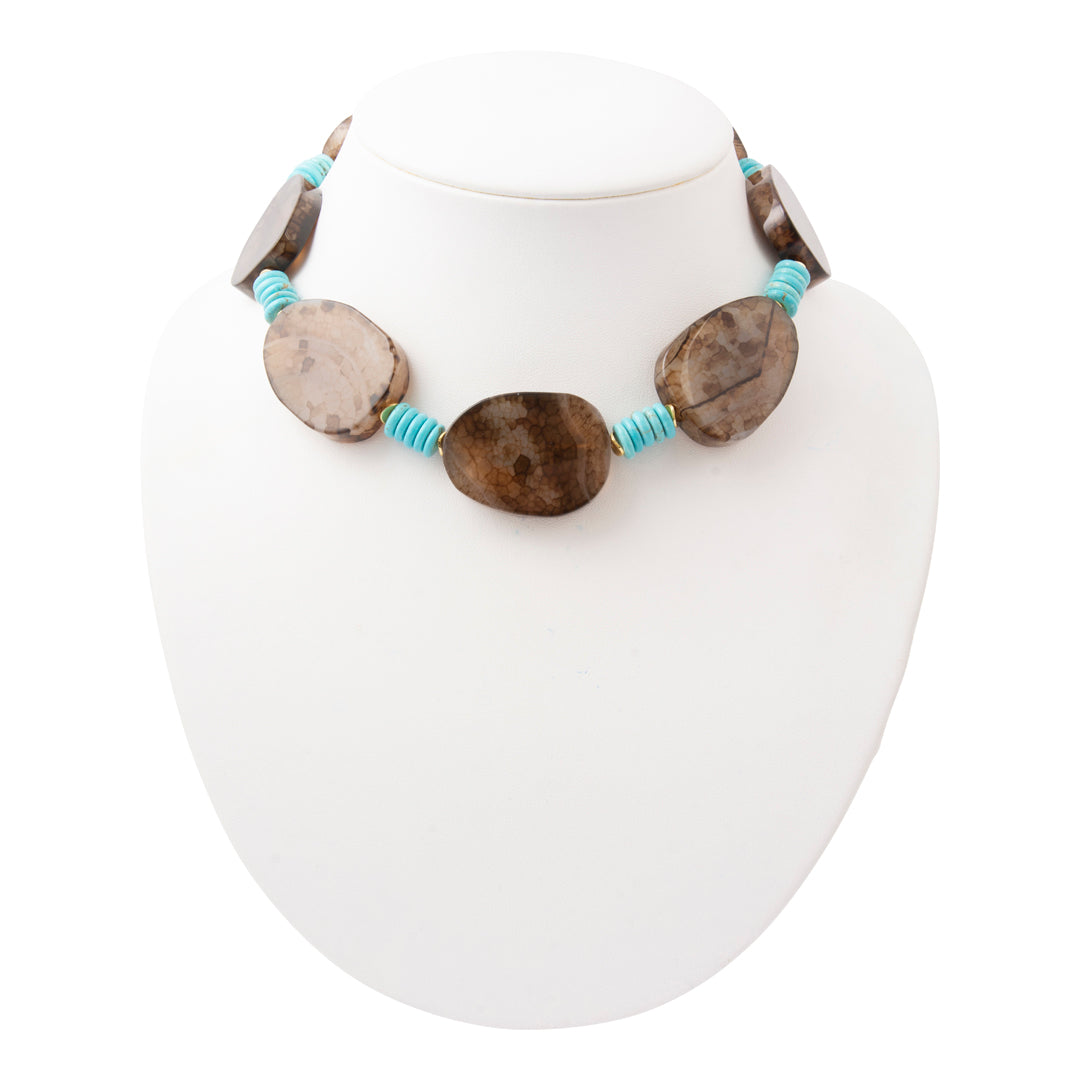 A statement necklace featuring polished natural stone oval beads in brown and beige, separated by bright turquoise blue cord spacers, displayed on a white mannequin bust.