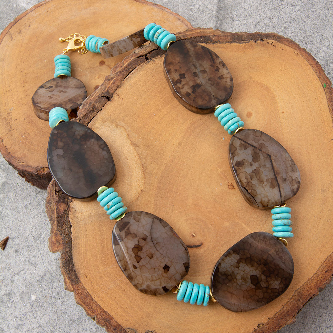 A beaded necklace with large polished brown stone pendants and turquoise spacer beads, displayed on a rustic wood slice over a gray surface.