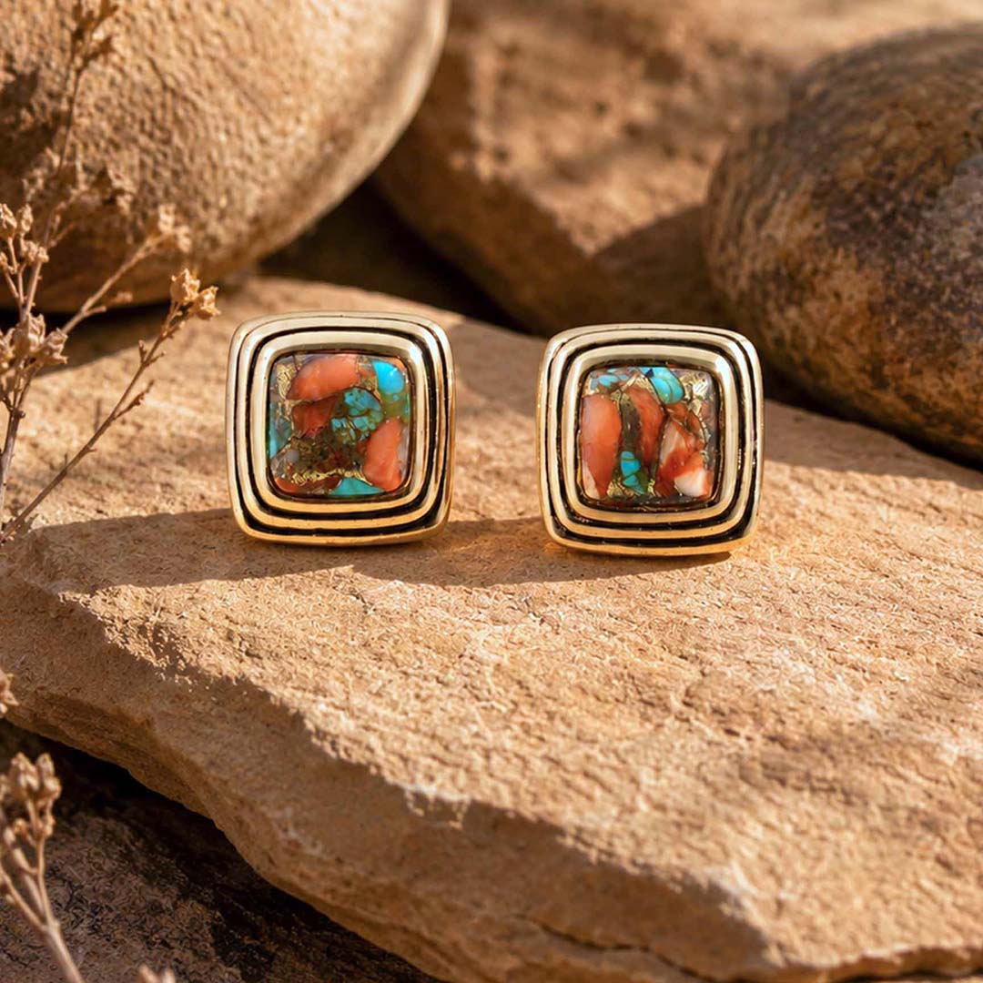 A pair of gold square stud earrings with a central inlay of orange and turquoise stone, displayed on a textured tan rock surface.