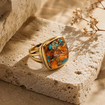 A gold ring with a square cabochon turquoise and orange stone, resting on a textured beige stone block with dried flowers in the background.