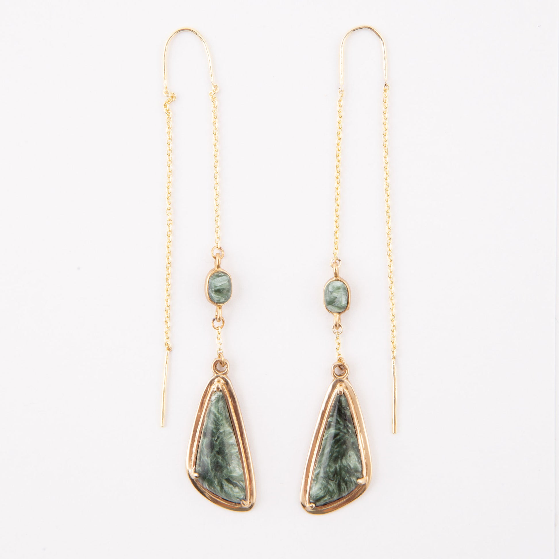 A pair of gold drop earrings featuring a small round green stone and a larger teardrop-shaped green stone with a gold border, hanging from delicate gold chains on a white background.