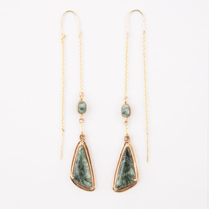A pair of gold drop earrings featuring a small round green stone and a larger teardrop-shaped green stone with a gold border, hanging from delicate gold chains on a white background.