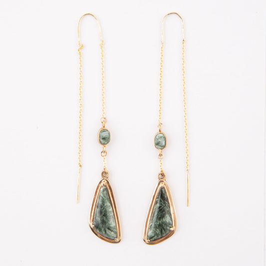 A pair of gold drop earrings featuring a small round green stone and a larger teardrop-shaped green stone with a gold border, hanging from delicate gold chains on a white background.