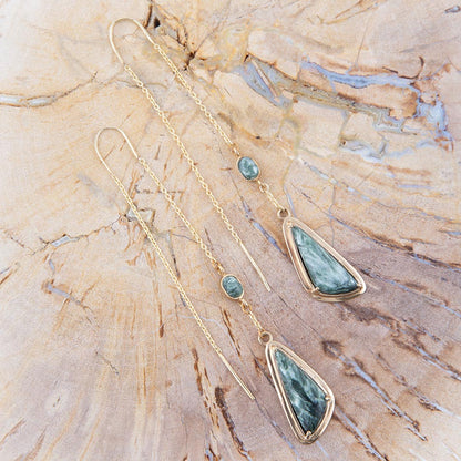 A pair of gold dangle earrings with triangular green stone pendants and small round green stones, displayed on a textured wood surface.