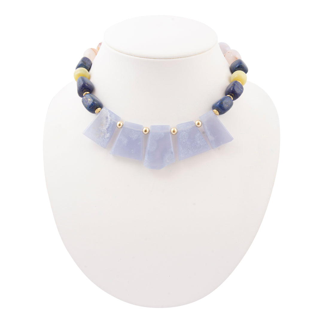 A beaded necklace displayed on a white jewelry bust. The necklace features a central row of pale lavender-blue stone pieces separated by small gold beads, with dark blue and cream-colored round beads along the upper chain.