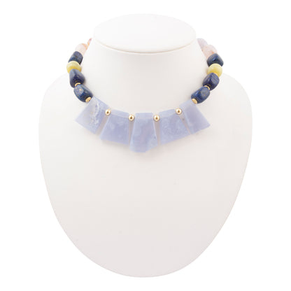 A beaded necklace displayed on a white jewelry bust. The necklace features a central row of pale lavender-blue stone pieces separated by small gold beads, with dark blue and cream-colored round beads along the upper chain.