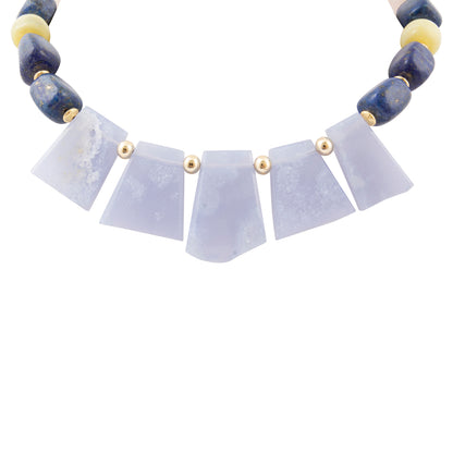 A beaded necklace featuring five light lavender-gray agate fan-shaped beads in the center, flanked by dark blue lapis lazuli and cream-colored round beads separated by small gold spacers, set against a plain white background.