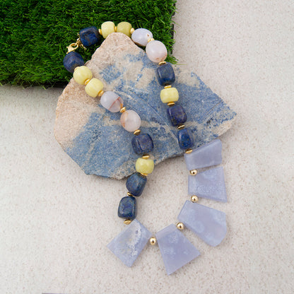 A beaded necklace with dark blue, pale yellow, and white beads, draped over a blue stone slab on a light textured surface with green moss in the corner.