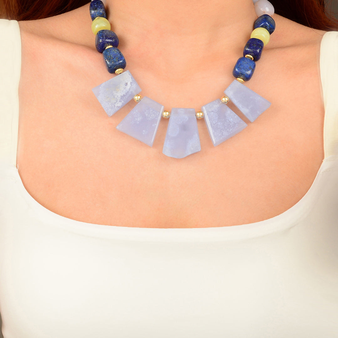 A close-up of a woman wearing a statement necklace with five light blue stone slices and blue, navy, and pale yellow beads, paired with a white scoop-neck top.