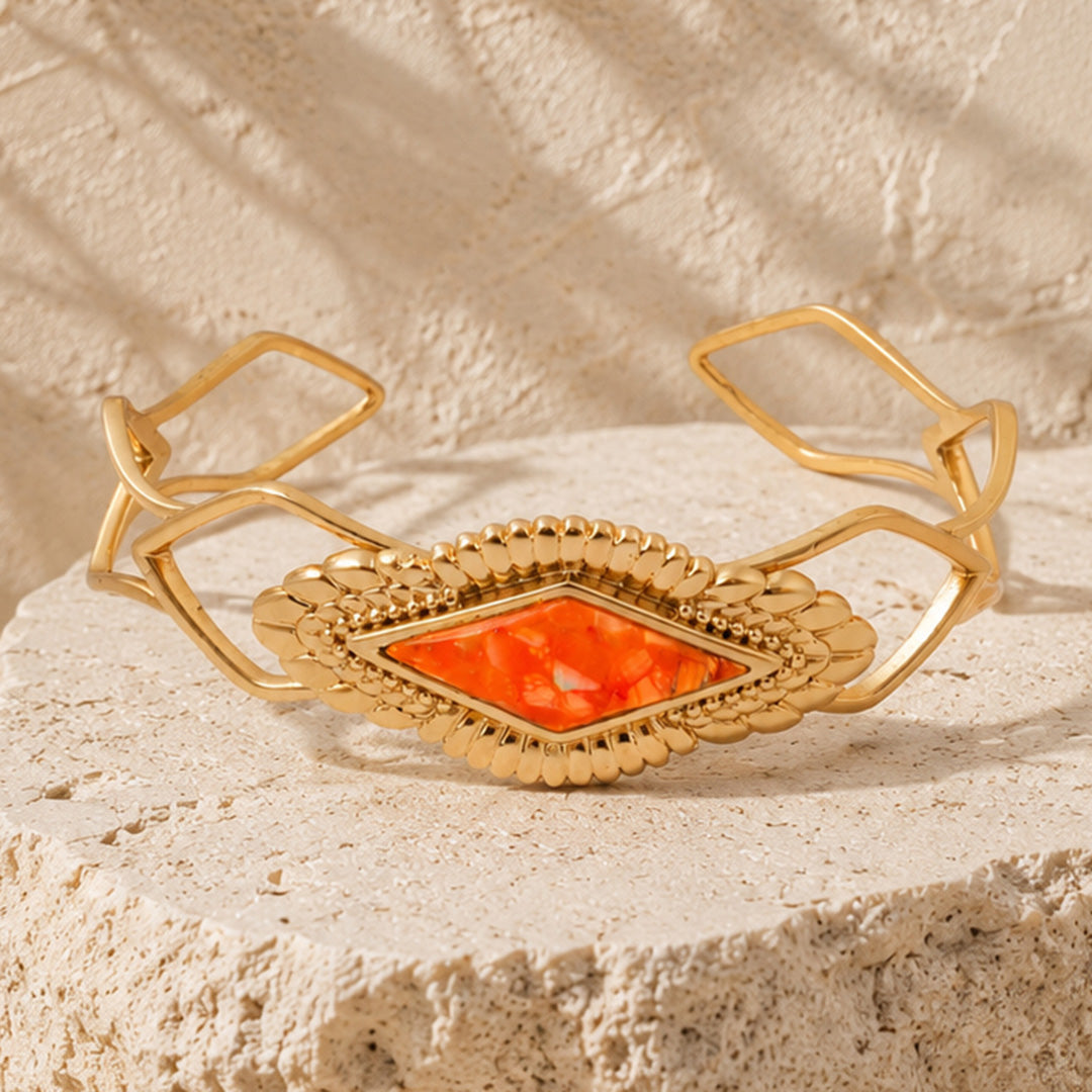 A gold-toned open cuff bracelet with a textured central diamond-shaped orange stone, displayed on a light beige stone surface.