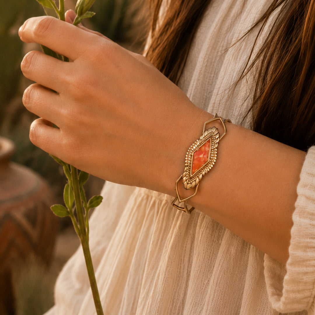 A gold-toned cuff bracelet with a central peach-orange stone, worn on a woman's wrist. The woman is wearing a cream-colored textured dress and holding a small green sprig, with a warm, blurred outdoor background.