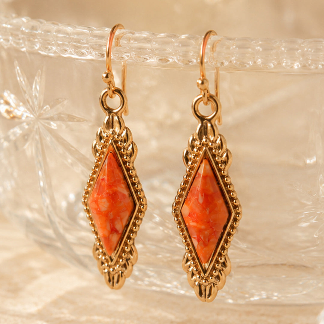 A pair of gold-tone drop earrings with diamond-shaped orange and white marbled stone centers, displayed on a textured clear glass surface.