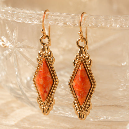 A pair of gold-tone drop earrings with diamond-shaped orange and white marbled stone centers, displayed on a textured clear glass surface.