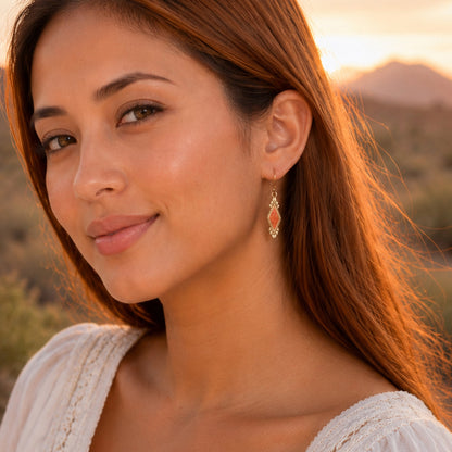 A close-up portrait of a woman outdoors at sunset wearing a dangling gold earring with a coral-pink stone, against a blurred natural landscape background.