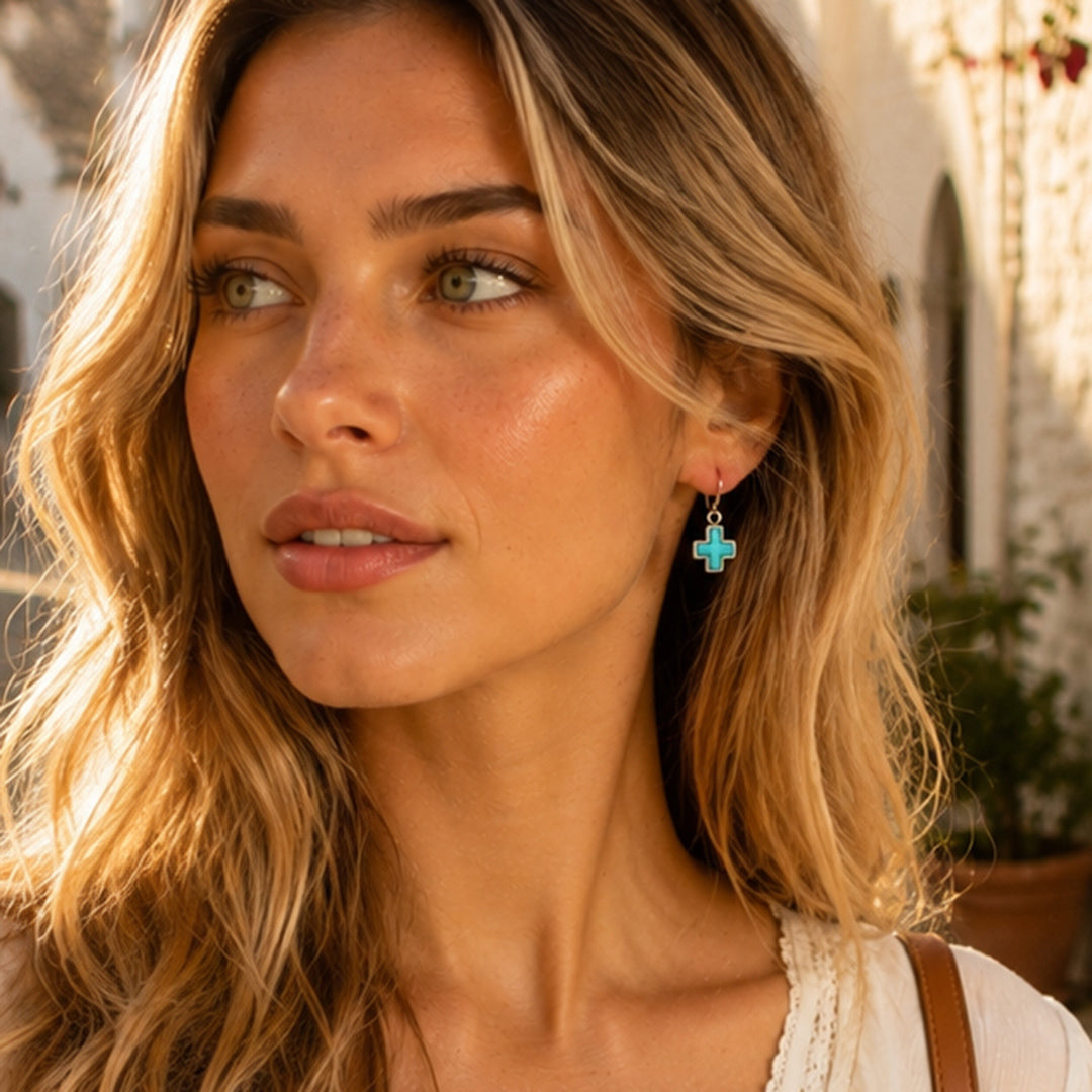 A close-up lifestyle shot of a woman with blonde hair wearing a small turquoise cross dangle earring with a gold-tone hook.