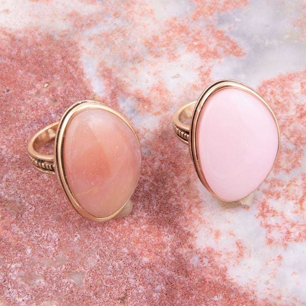 Two gold rings with large oval pink cabochon gemstones, displayed on a textured pink and white stone surface.