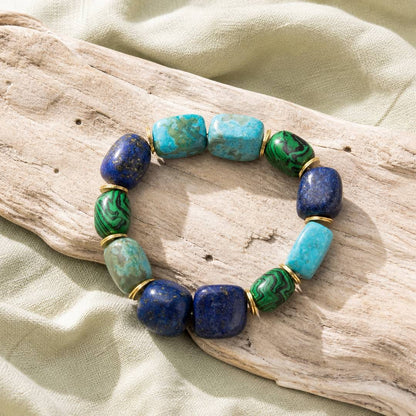 A beaded stretch bracelet with blue lapis lazuli, turquoise, and green malachite beads separated by gold spacer rings, displayed on a piece of driftwood over a light green fabric background.