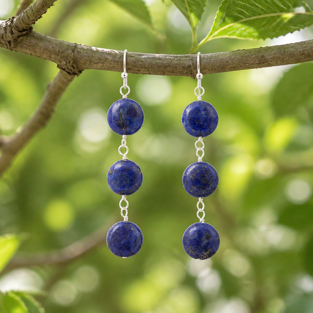 A pair of silver drop earrings with four deep blue lapis lazuli sphere beads hanging from a tree branch with green leaves in the background.
