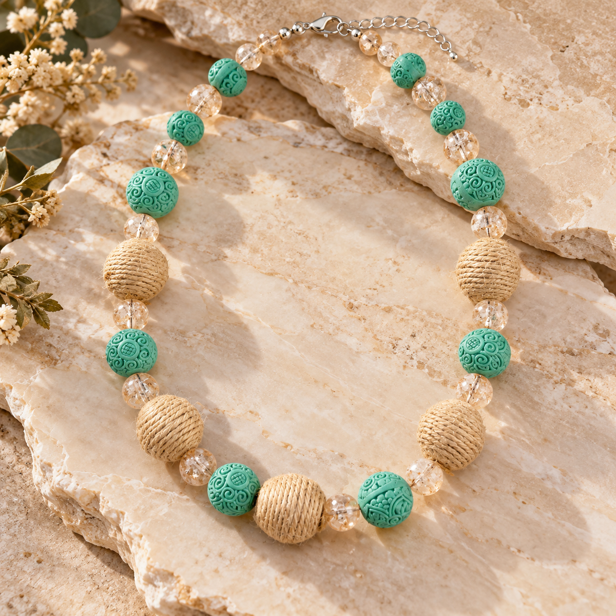 A beaded necklace with turquoise green carved round beads, natural tan wrapped beads, and clear faceted crystal beads, displayed on beige stone slabs with small dried flowers.