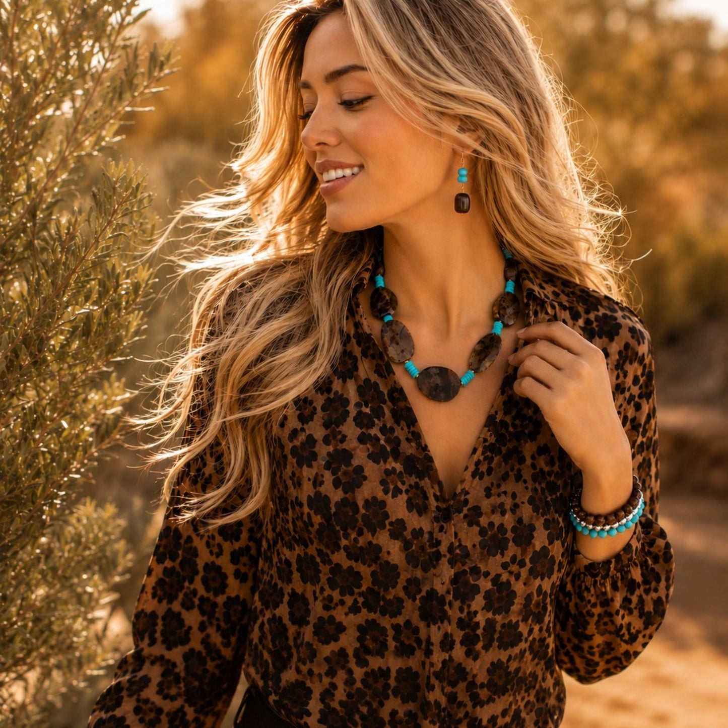 A woman outdoors wearing a leopard-print blouse and turquoise-and-black statement jewelry, including a necklace and bracelet, with warm golden sunlight and blurred foliage in the background.