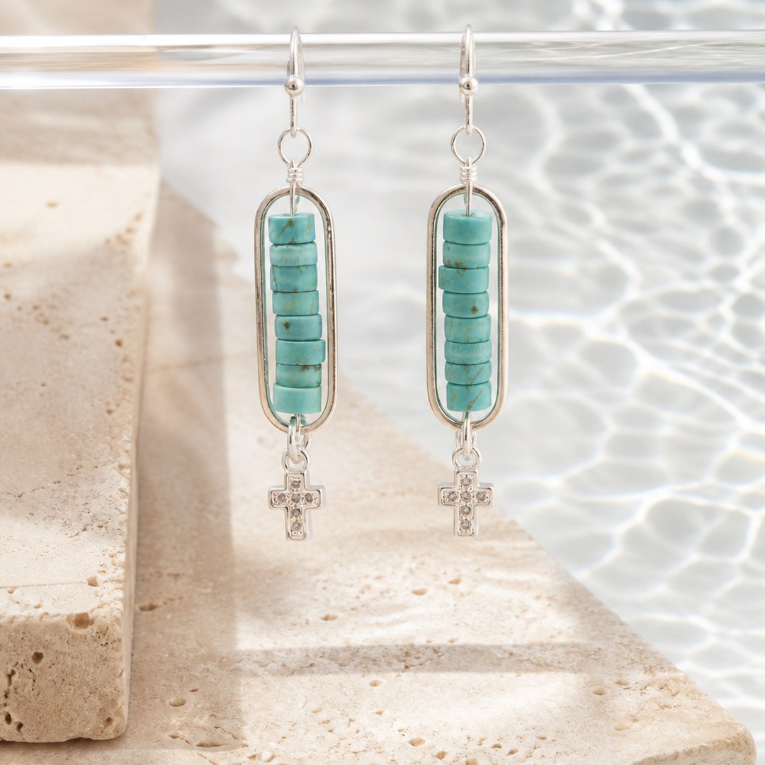 A pair of silver dangle earrings with turquoise stone beads and small cross charms, displayed on a clear stand over a stone surface with water in the background.