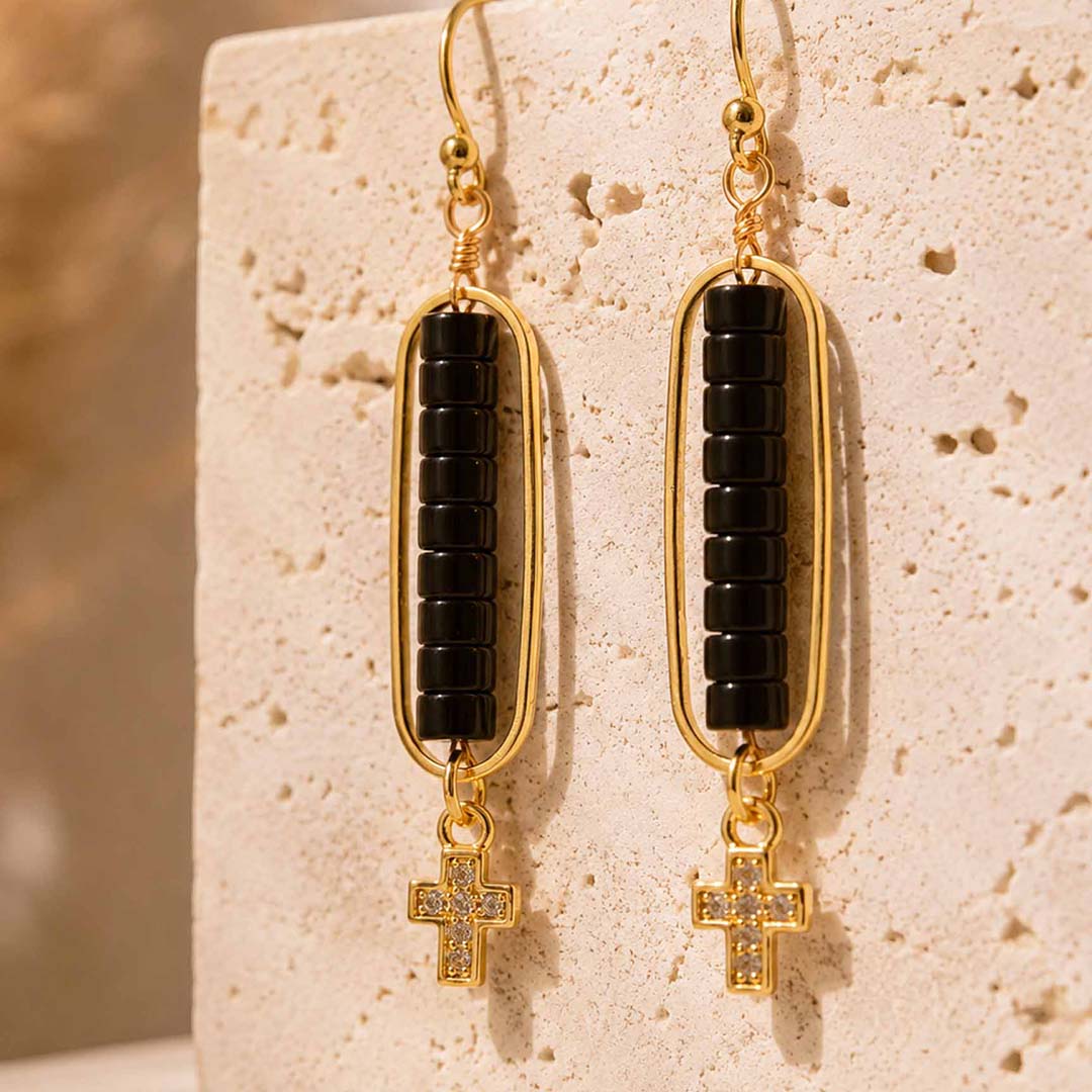 A pair of gold drop earrings with black beaded rectangular bars and small cross charms, displayed against a textured beige stone block.