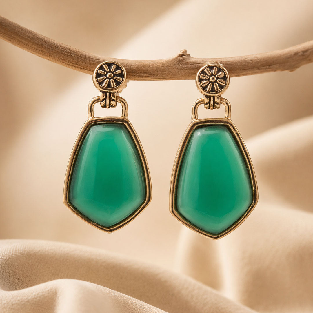 A pair of gold-tone drop earrings with large, smooth green teardrop-shaped stones, hanging from a natural branch against a soft beige fabric background.