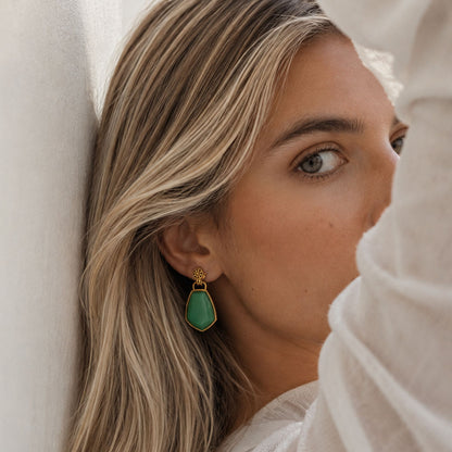 A close-up lifestyle shot of a woman wearing a gold drop earring with a green teardrop-shaped stone, leaning against a white textured wall.