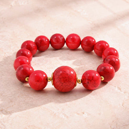 A beaded stretch bracelet made of glossy red marbled round beads, featuring a larger central bead and small gold spacer beads, displayed on a light beige textured stone surface.