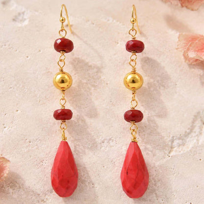 A pair of gold and red drop earrings with faceted red beads, polished gold spacer beads, and large teardrop-shaped red pendants, displayed on a textured light beige surface.