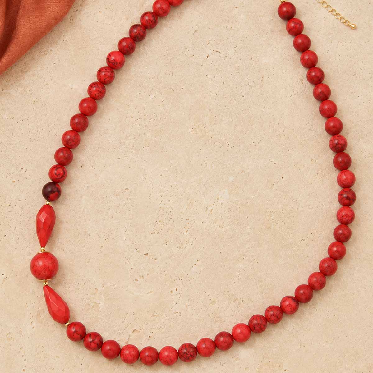 A red beaded necklace with a mix of matte and polished round beads, a faceted red teardrop bead, and a small gold clasp, displayed on a textured beige surface.