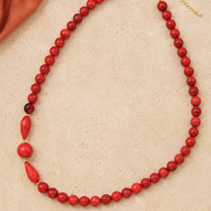 A red beaded necklace with a mix of matte and polished round beads, a faceted red teardrop bead, and a small gold clasp, displayed on a textured beige surface.