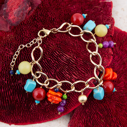A gold-tone chain bracelet with colorful faux-stone beads and dangling charms, arranged on a textured deep red fabric background.
