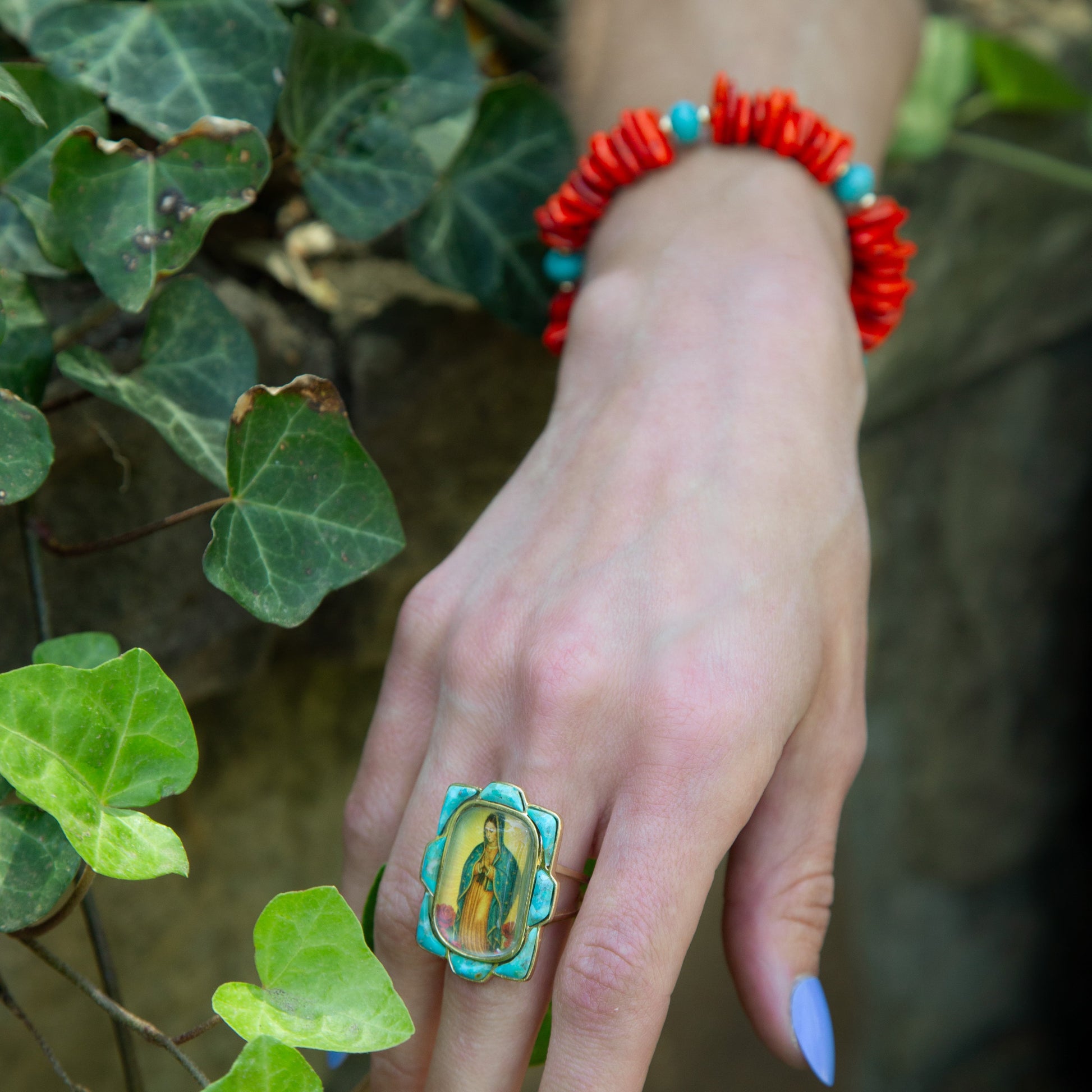 Hand wearing a turquoise ring and red bracelet with green leaves in the background