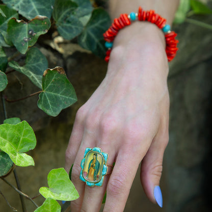 Hand wearing a turquoise ring with a Guadalupe Virgin design, surrounded by green leaves.