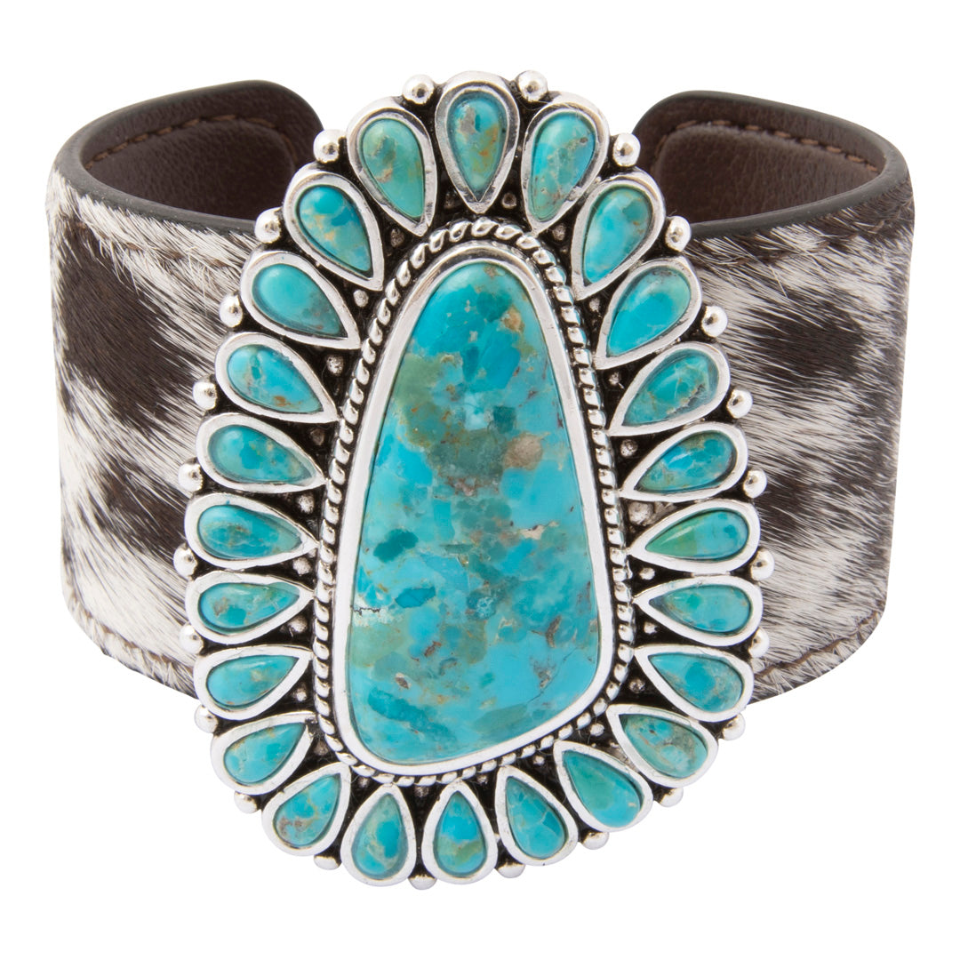 A wide cuff bracelet featuring a large central turquoise stone surrounded by smaller turquoise stones in a silver-tone border, with a black-and-white cowhide strap in the background.
