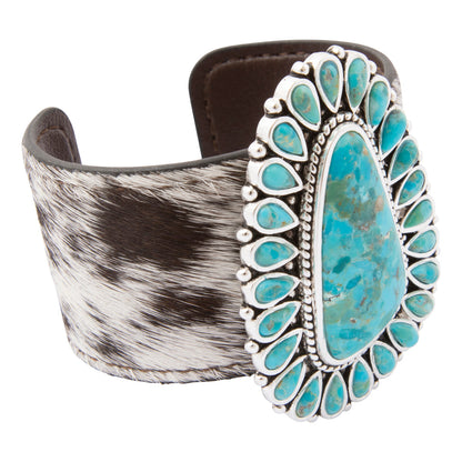 A wide cuff bracelet featuring a large turquoise stone centerpiece surrounded by a border of small turquoise stones and silver-tone metal, with a wide brown and black cowhide-textured band on the left side.