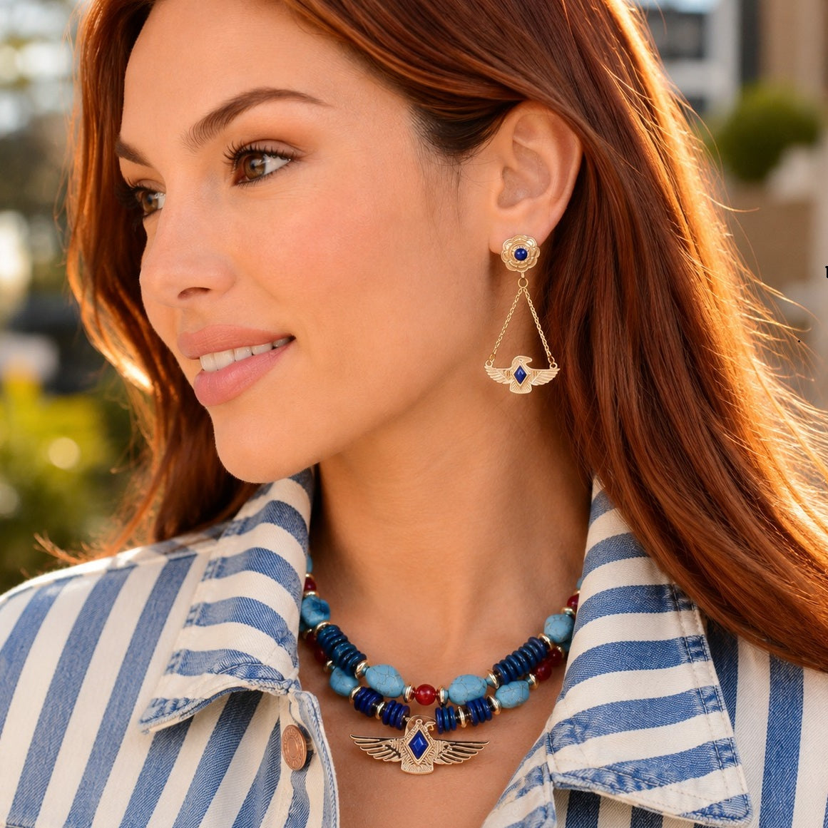 A woman wearing a matching gold-toned winged earring and necklace set with blue and red details, paired with a blue and white striped shirt.