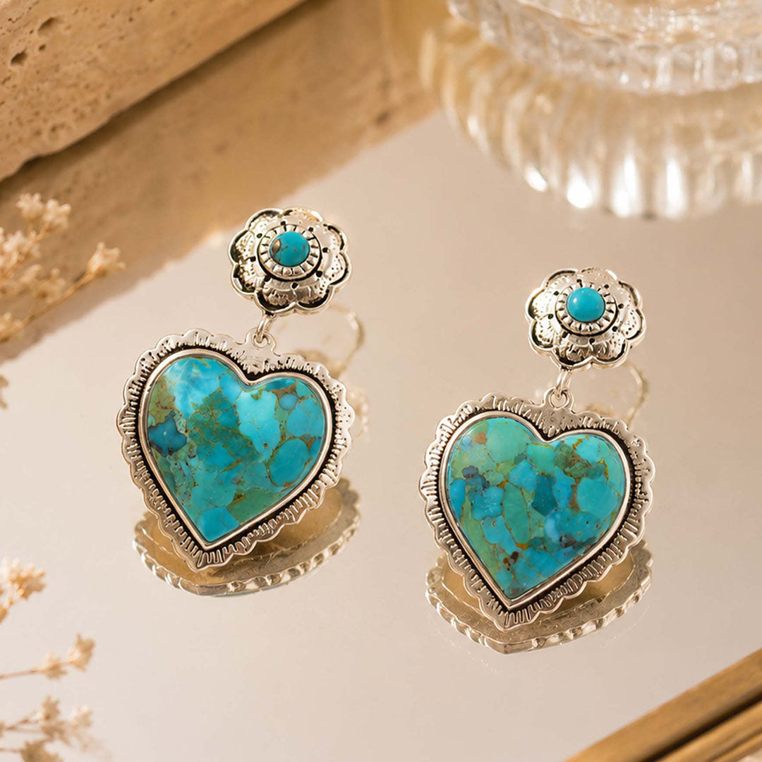 A pair of silver-toned drop earrings with heart-shaped turquoise stones and small round turquoise accents at the top, displayed on a reflective surface with a warm neutral background.