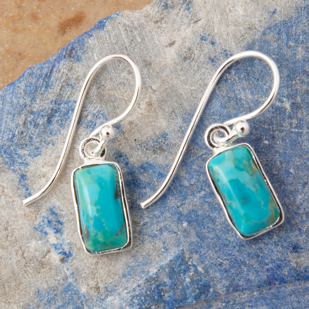 A pair of silver drop earrings with rectangular turquoise-blue stone pendants, displayed on a textured blue and gray stone surface.