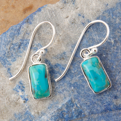 A pair of silver drop earrings with rectangular turquoise-blue stone pendants, displayed on a textured blue and gray stone surface.