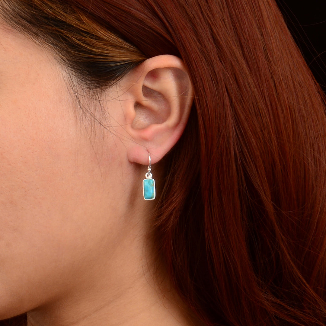 A close-up of a person's ear wearing a silver dangle earring with a small rectangular light blue stone, against a dark background.