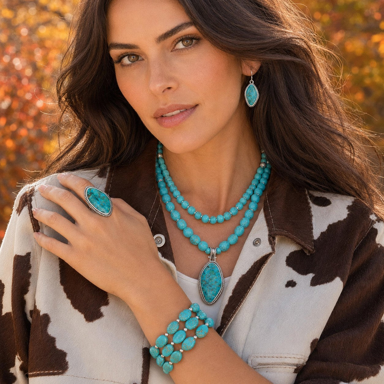 A woman wearing a matching turquoise jewelry set, including a necklace with a large oval pendant, a ring, and a bracelet, styled with a brown and white cow-print jacket against an autumnal outdoor background.